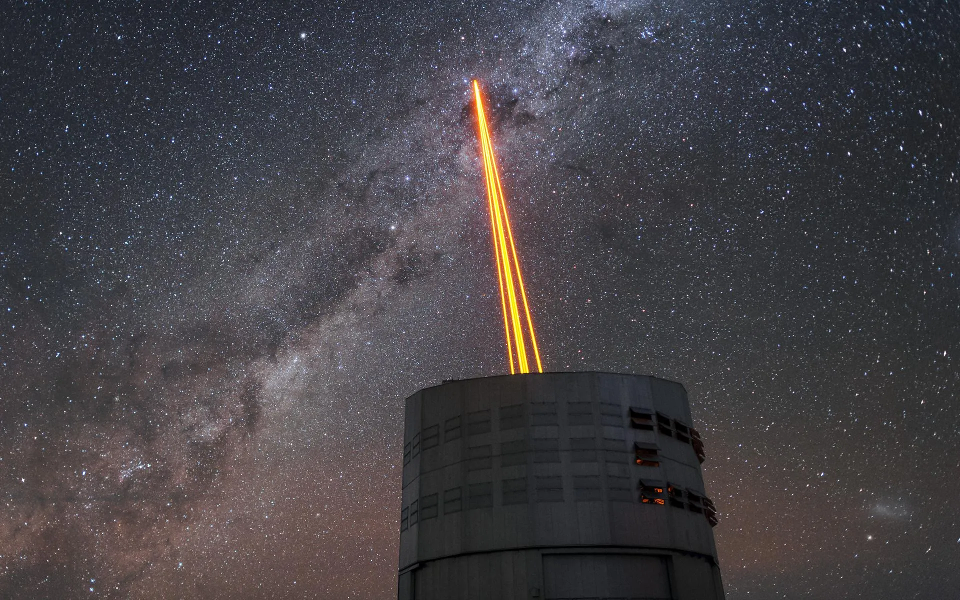 Four orange laser beams shine from a telescope dome into a night sky full of stars and the Milky Way.