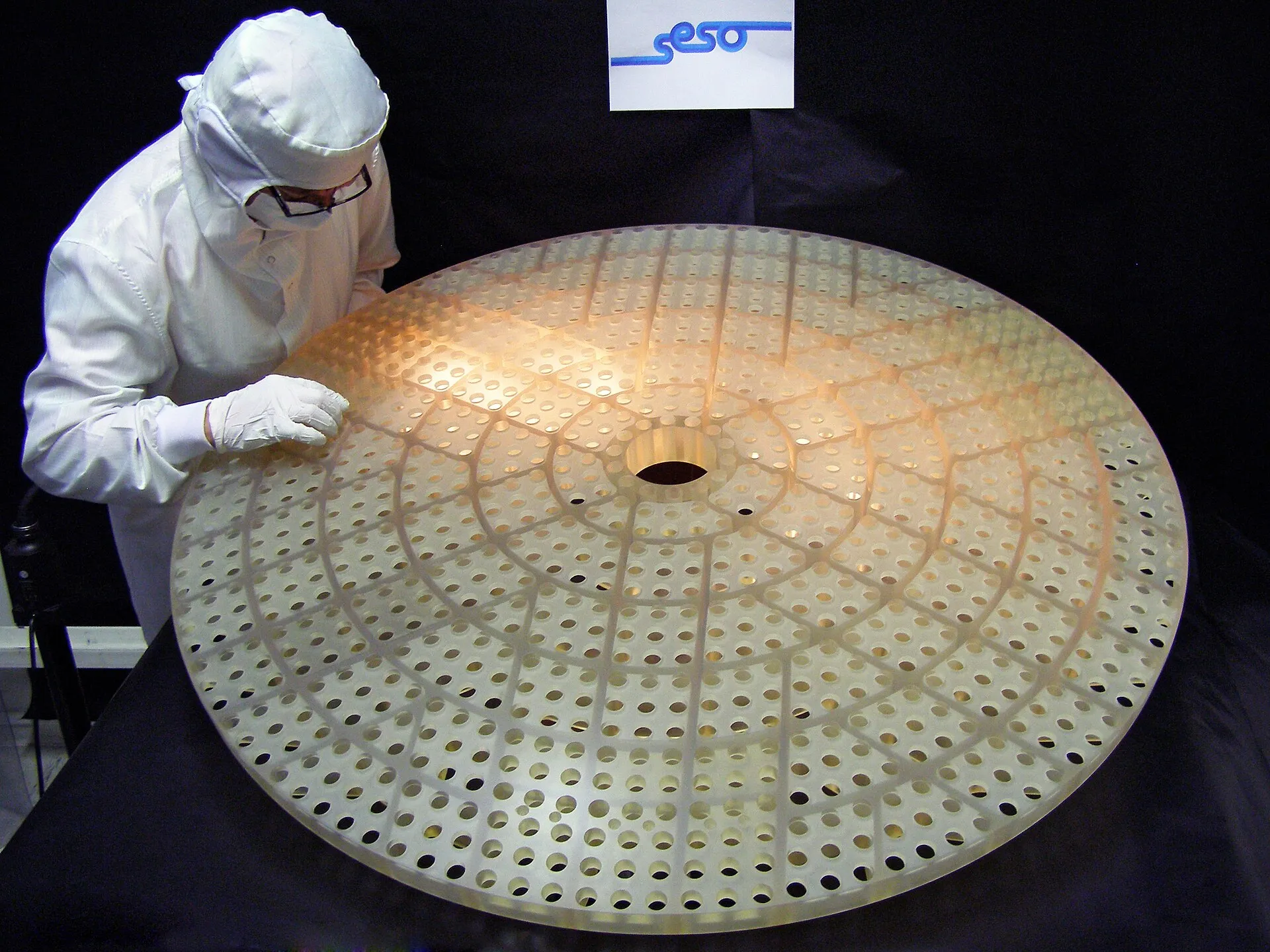 A technician in a clean-room suit leans over a large round disc pierced by hundreds of evenly spaced holes.
